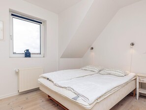 Une chambre avec un plafond en pente, un lit avec une literie blanche, un radiateur, une fenêtre donnant sur un beau paysage et une petite table de chevet.
