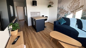 Una sala de estar moderna con una televisión de pantalla plana, un sofá azul, una mesa de centro de madera y una cocina con salpicadero de azulejos y una lámpara colgante.