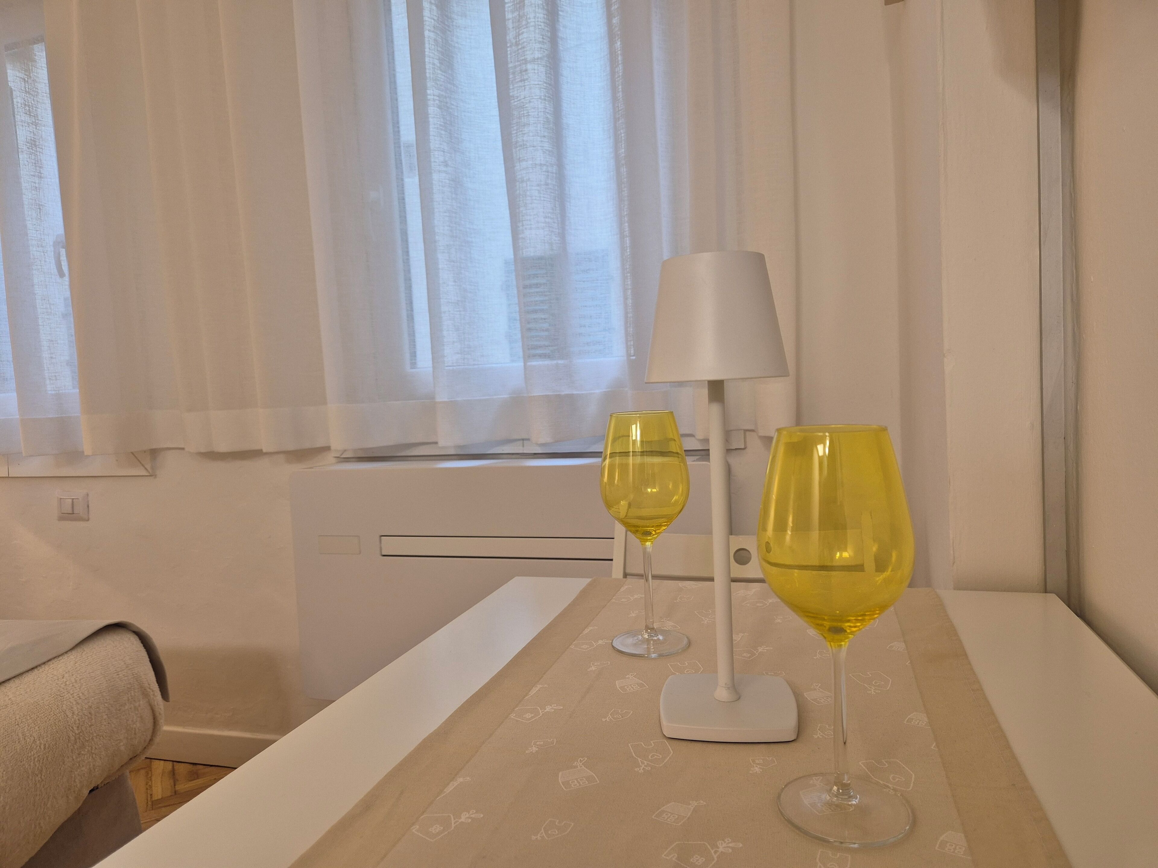 Une chambre d’hôtel avec un lit, une table de chevet sur laquelle reposent deux verres à vin, une lampe et une fenêtre avec des rideaux.