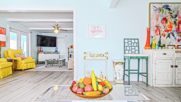 A living room with a glass coffee table, yellow chairs, a TV, and a painting on the wall.