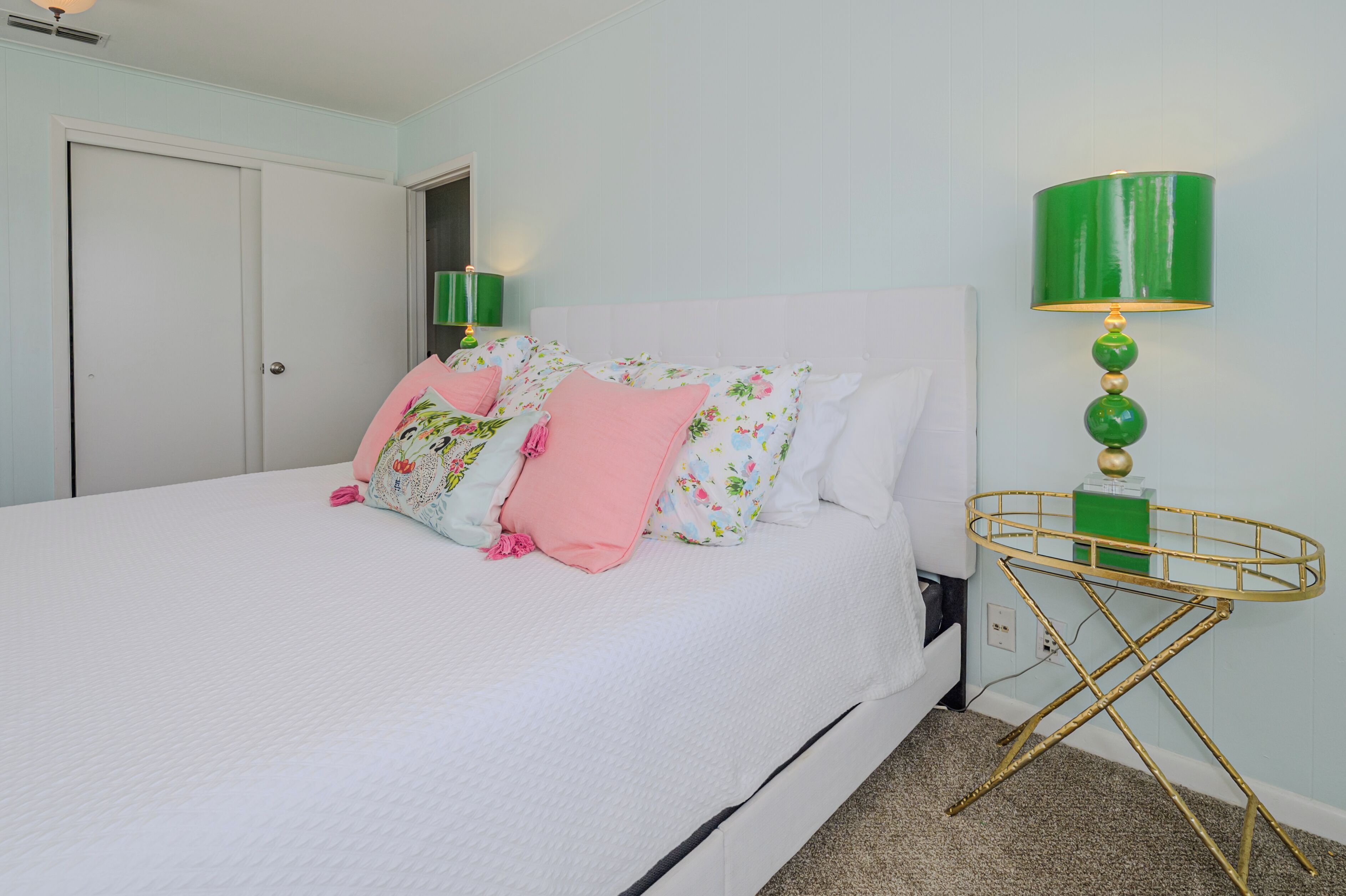 A neatly made bed with floral and pink pillows, a green lamp, and a gold-framed side table.