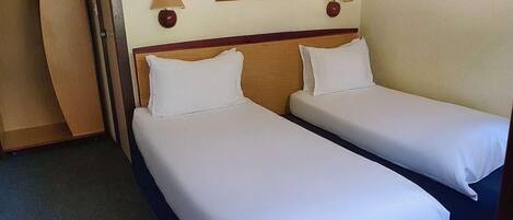 Quarto de hotel com duas camas de solteiro, cabeceira de madeira, quadro pendurado na parede e duas luminárias de cabeceira.