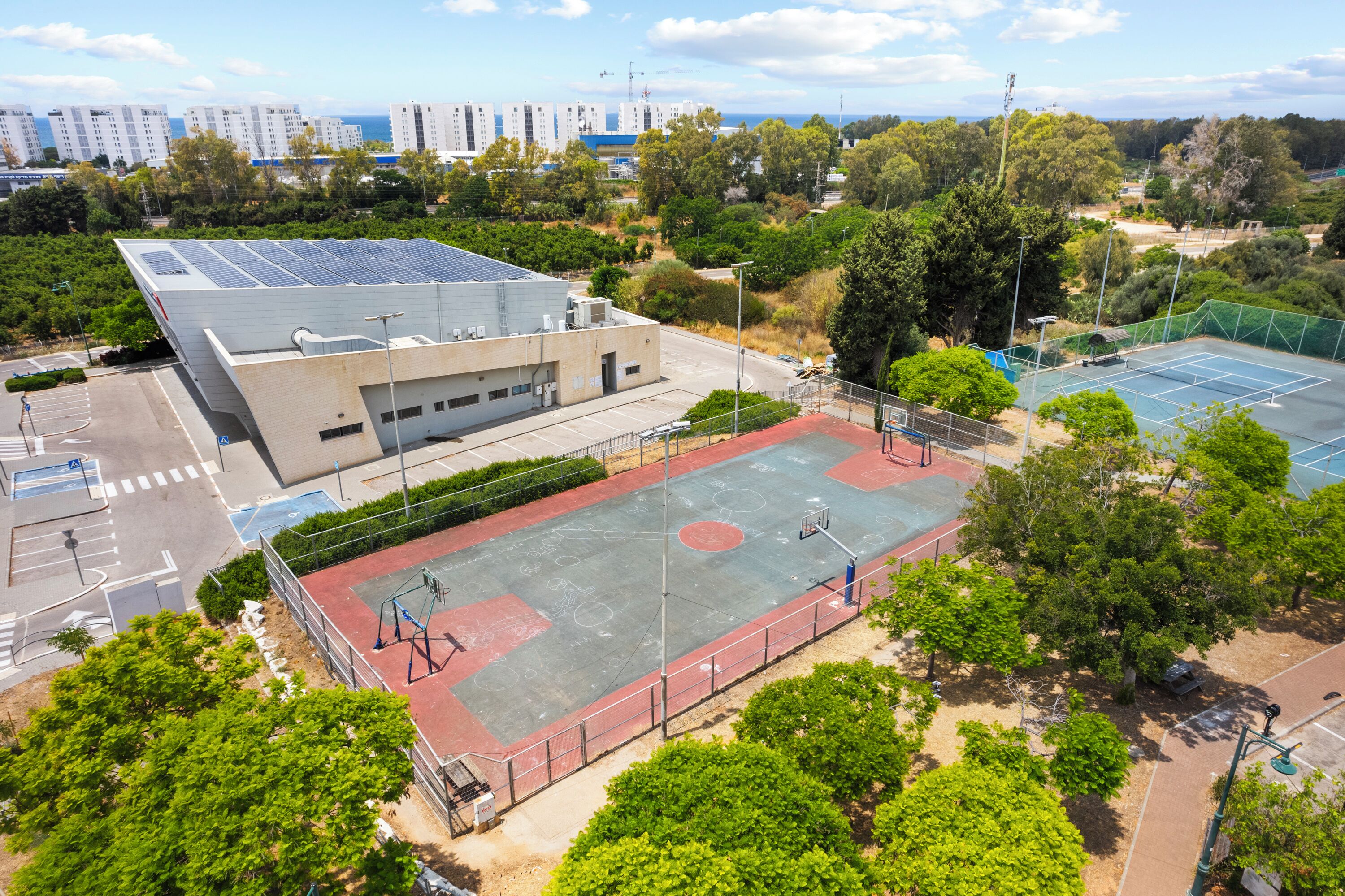 A sports court with a basketball hoop, surrounded by trees and a building with solar panels.