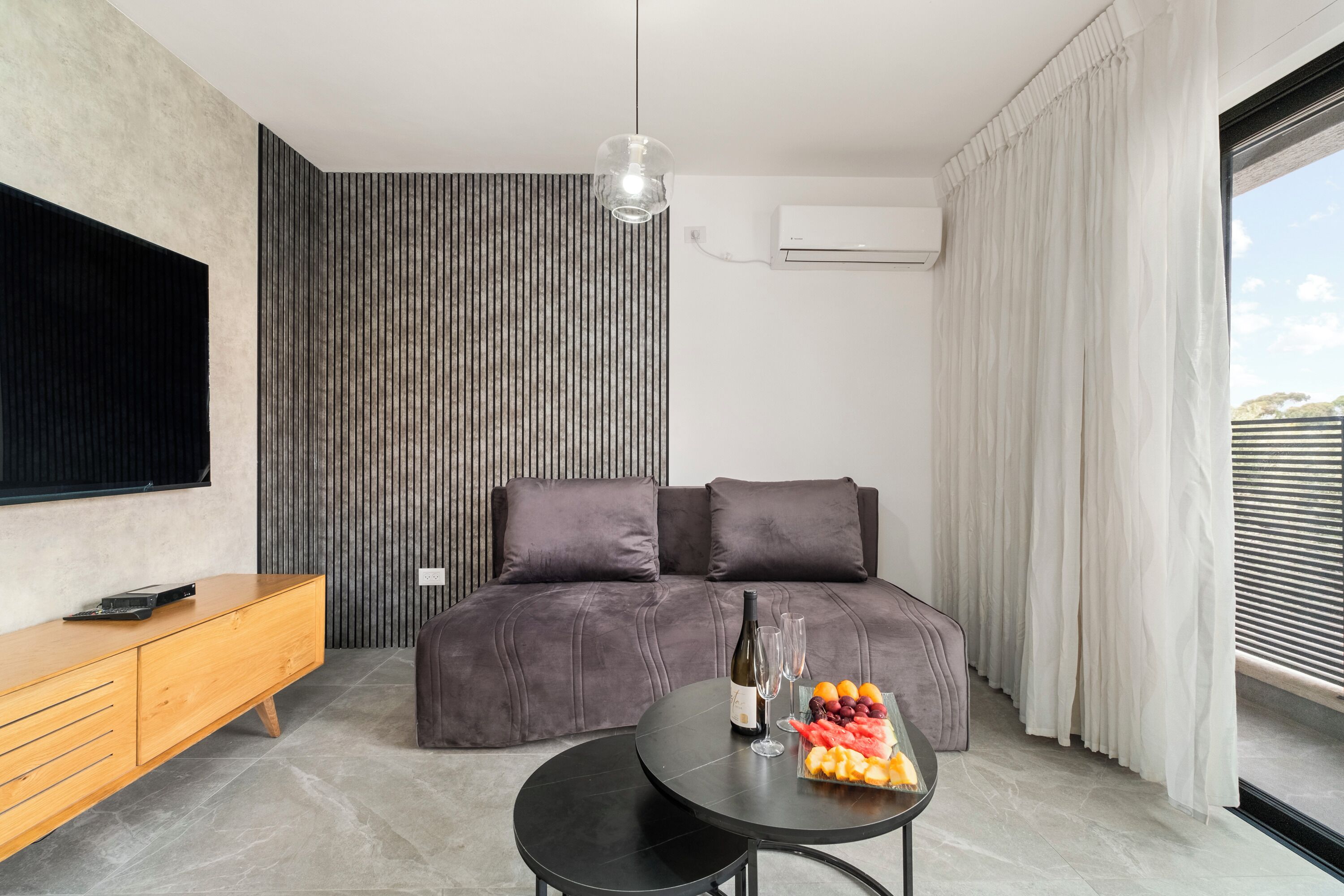 A modern bedroom with a sofa bed, a wooden side table, a round table with a bottle and glasses, and a fruit bowl.