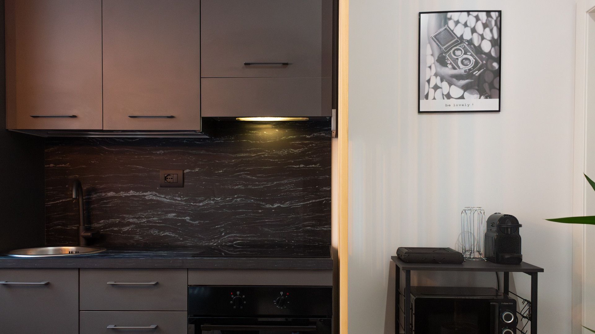 Cocina moderna con armarios oscuros, una salpicadura de mármol negro y una pequeña mesa negra con un altavoz y un vaso.