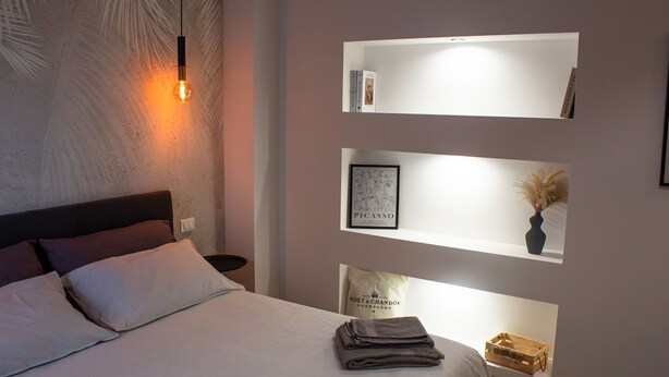 Une chambre à coucher moderne, comprenant un lit, une table de chevet, une étagère murale avec une œuvre d’art encadrée et un luminaire fixé au mur.