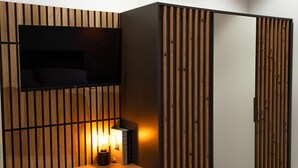 Une pièce avec un téléviseur fixé sur un mur recouvert de panneaux en bois, une armoire avec des lattes de bois verticales et une petite table avec une lampe.