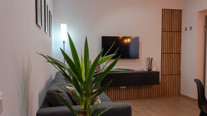 Un salon moderne avec un canapé gris, une grande plante en pot, un téléviseur à écran plat fixé au mur et un mur recouvert de panneaux en bois.