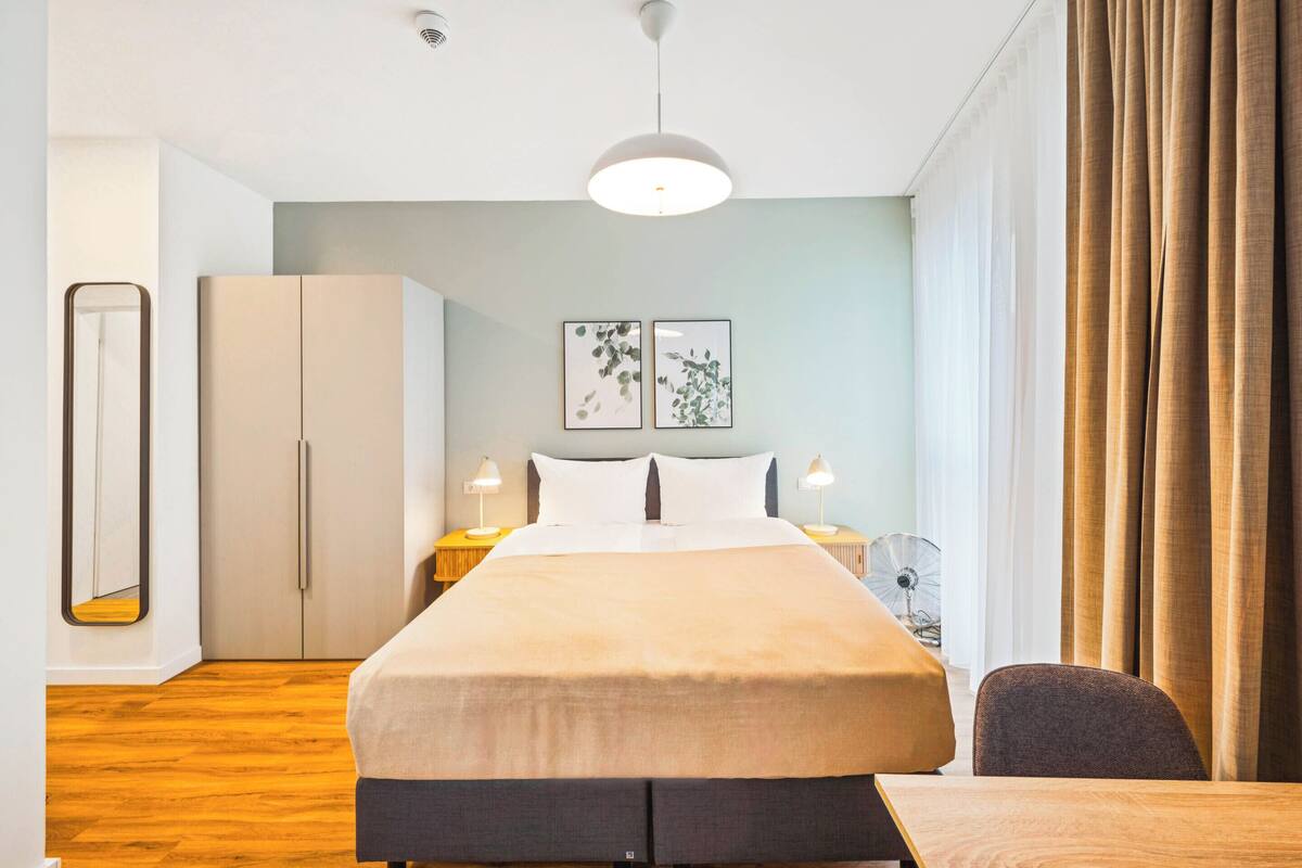 Una habitación de hotel moderna con una cama grande, mesitas de noche con lámparas, un armario y vistas al exterior.