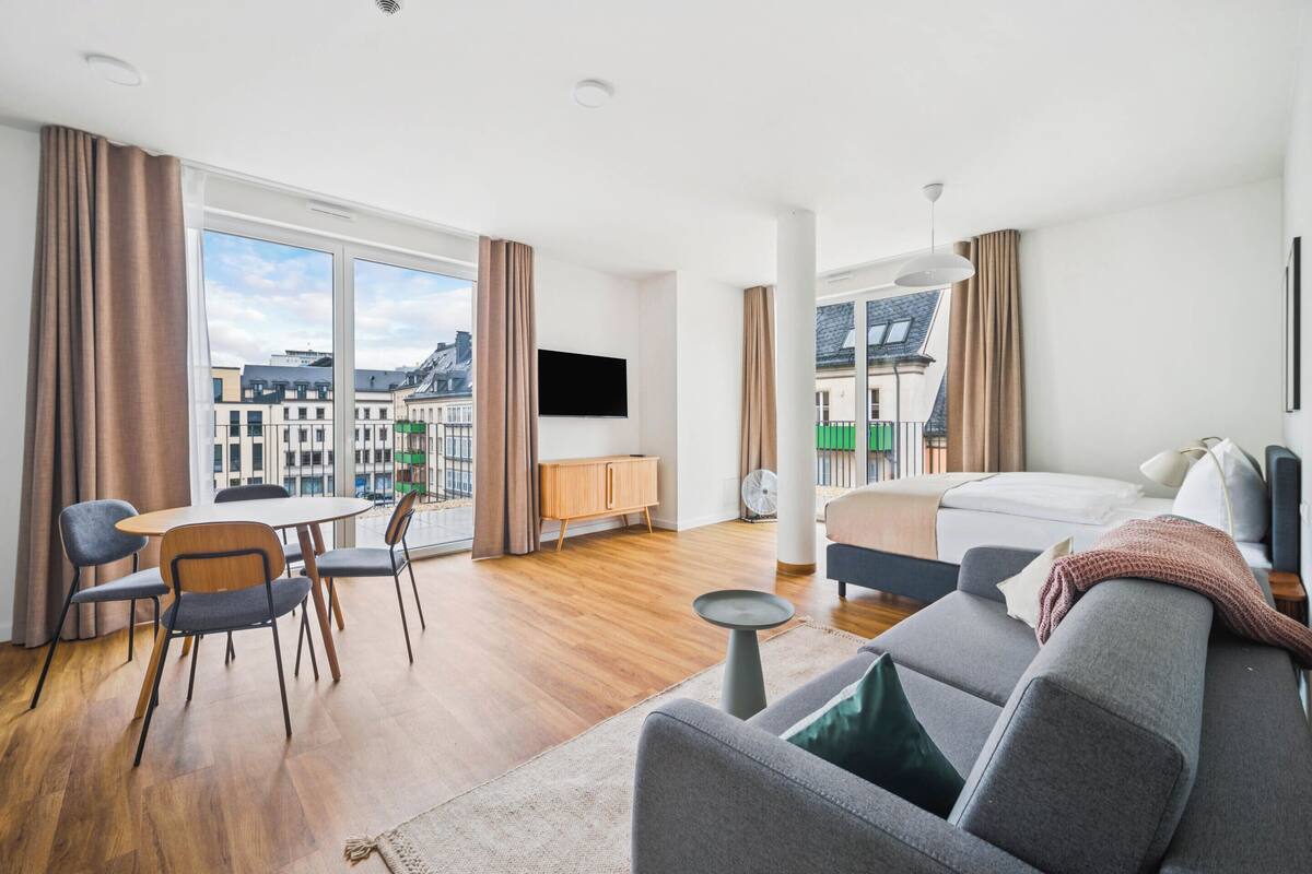 Una habitación de hotel moderna con un gran ventanal, un televisor de pantalla plana, un sofá, una mesa de comedor con sillas y una mesita auxiliar.