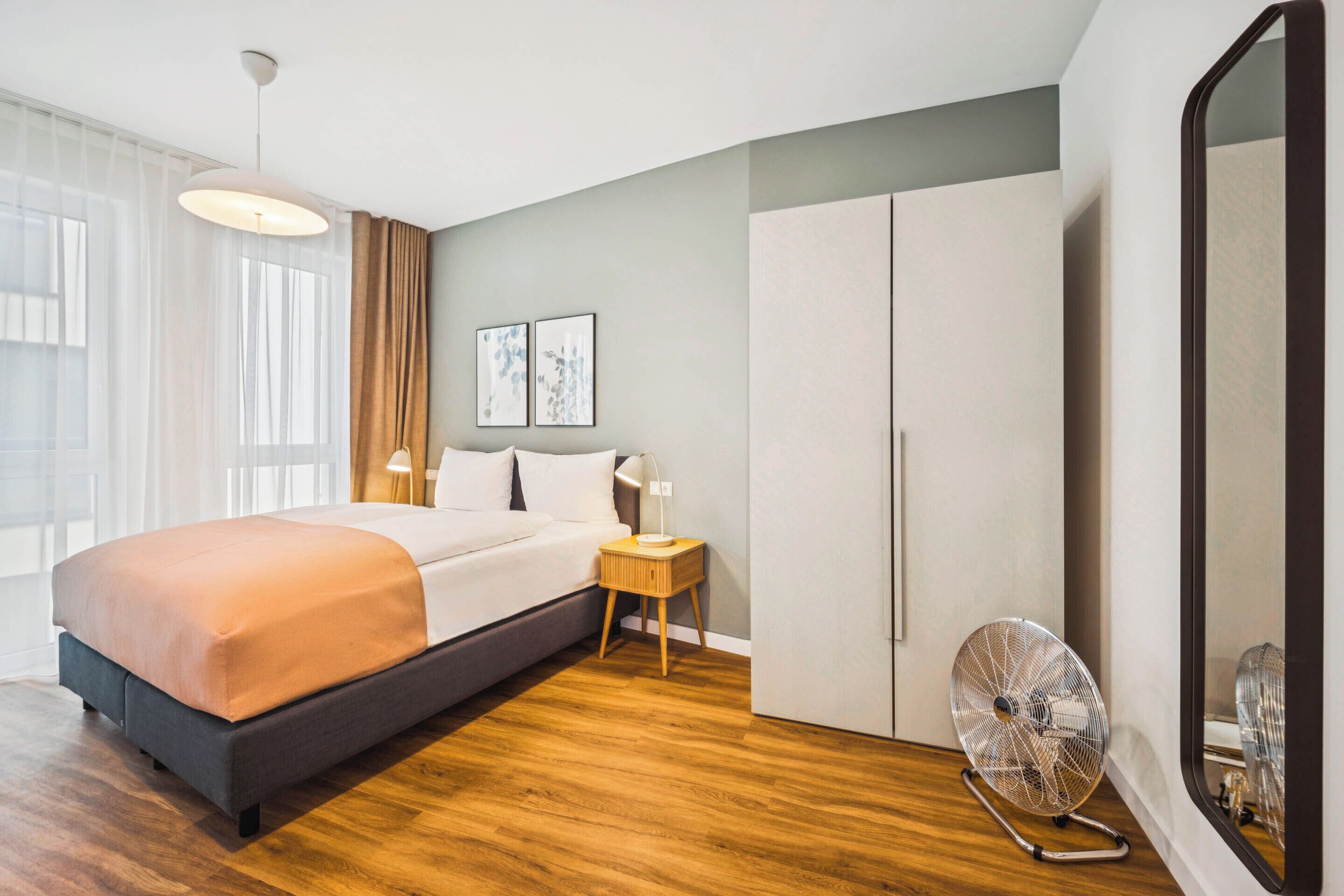 Une chambre moderne avec un grand lit, une table de chevet avec une lampe, un ventilateur sur pied et une armoire.