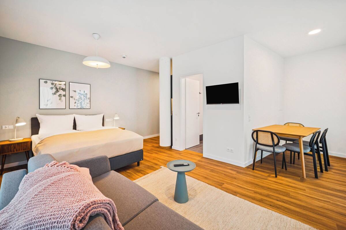 Una habitación de hotel moderna con una cama, un sofá, una mesa de comedor con sillas, un televisor montado en la pared y una mesita con un control remoto.