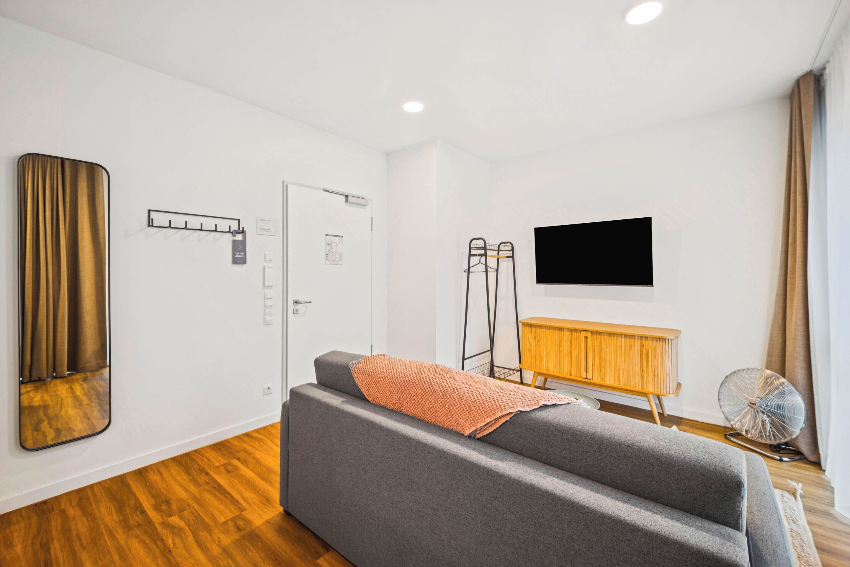 A modern hotel room with a grey sofa, a wooden cabinet, a flat-screen TV, a standing fan, and a large mirror.