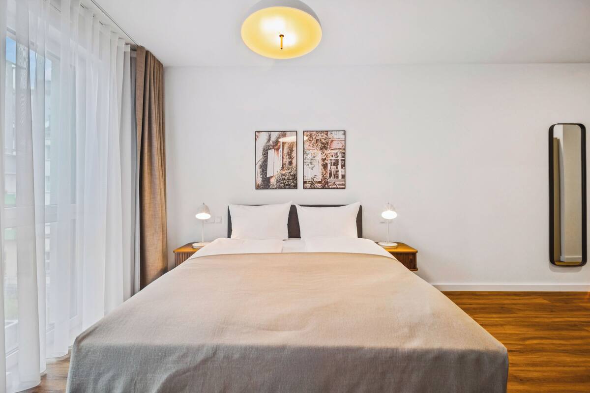 Una cama bien hecha con un edredón de tonos neutros, dos lámparas de noche y un espejo grande en la pared.