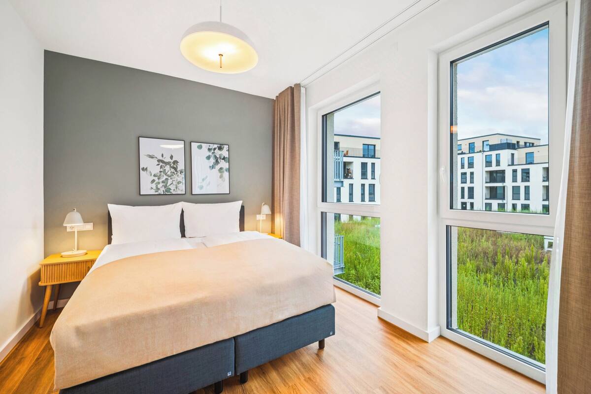 Un dormitorio moderno con una cama grande, dos mesitas de noche y un amplio ventanal que ofrece vistas a edificios y zonas verdes.