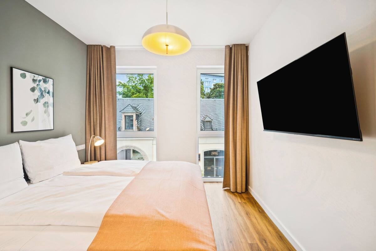 Habitación de hotel con cama, televisor de pantalla plana montado en la pared, ventana con cortinas y lámpara colgante.