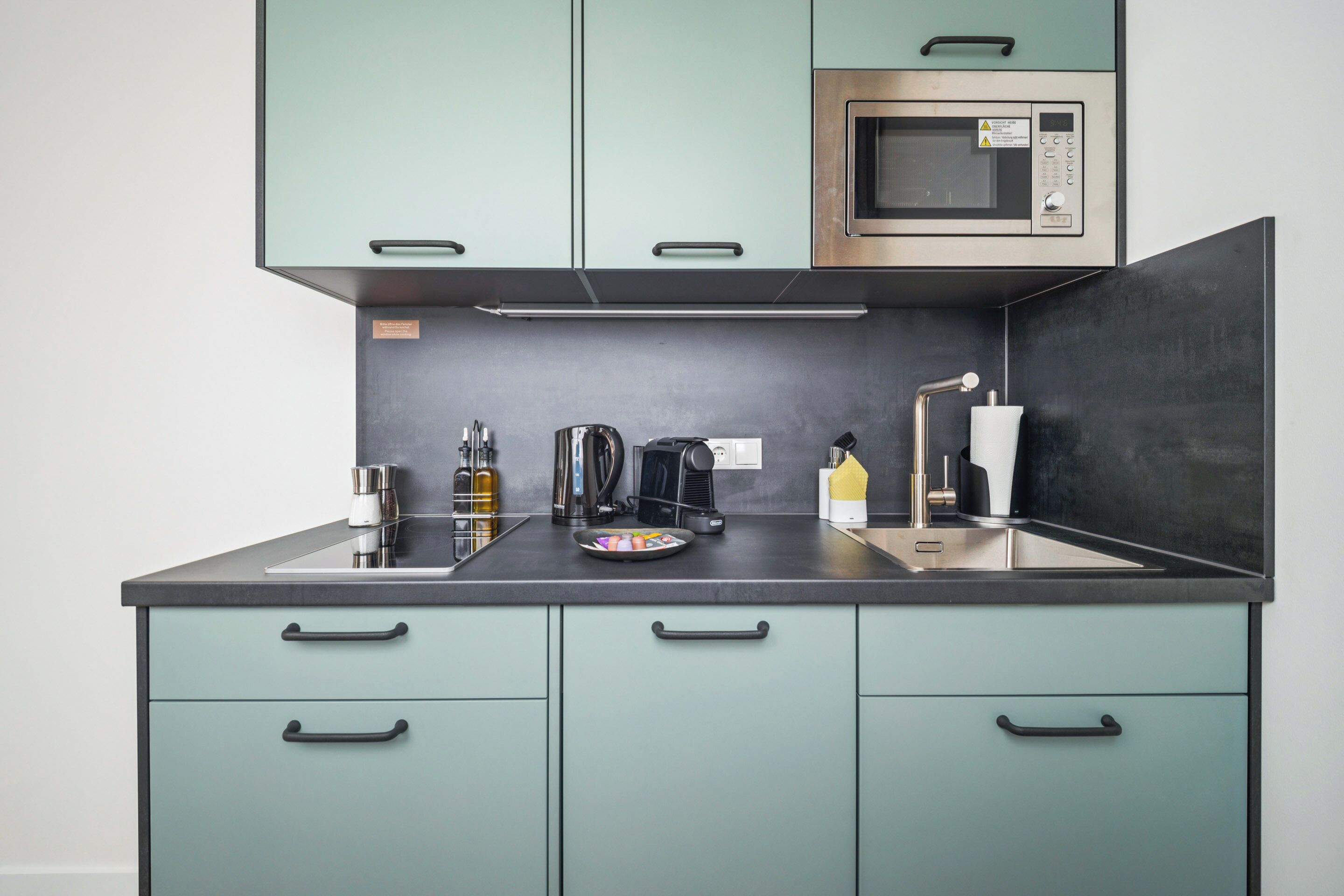 A compact kitchen with teal cabinets, a stainless steel microwave, a coffee maker, and a sink.