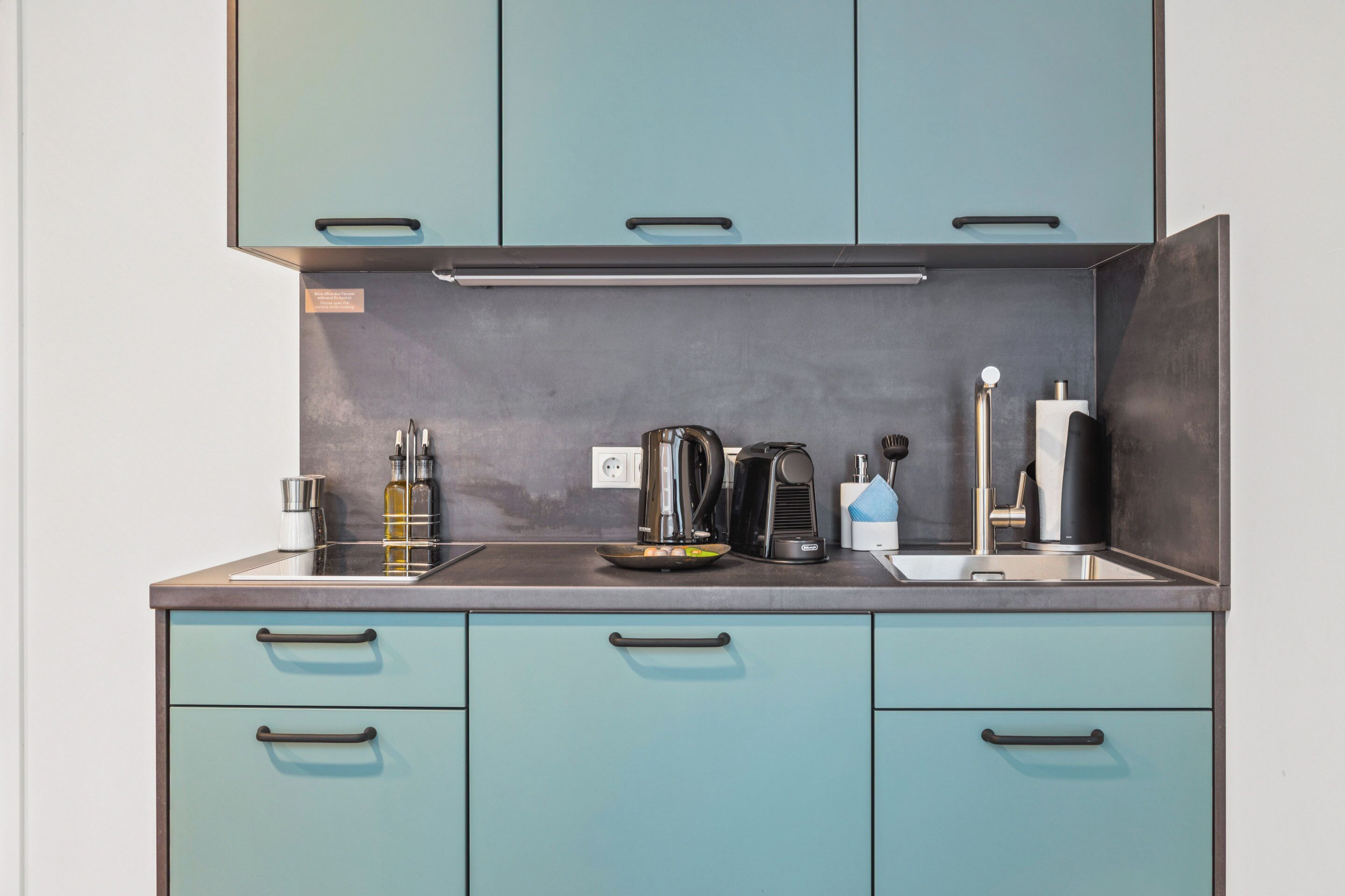 A kitchen with teal cabinets, a stainless steel sink, and modern appliances including a kettle and a coffee maker.