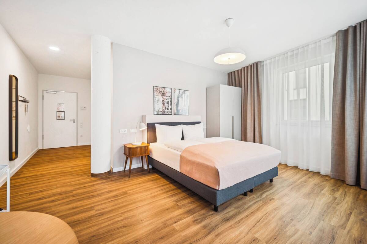 Habitación de hotel moderna con una cama grande, piso de madera, una mesita de noche con lámpara y un ventanal con cortinas.