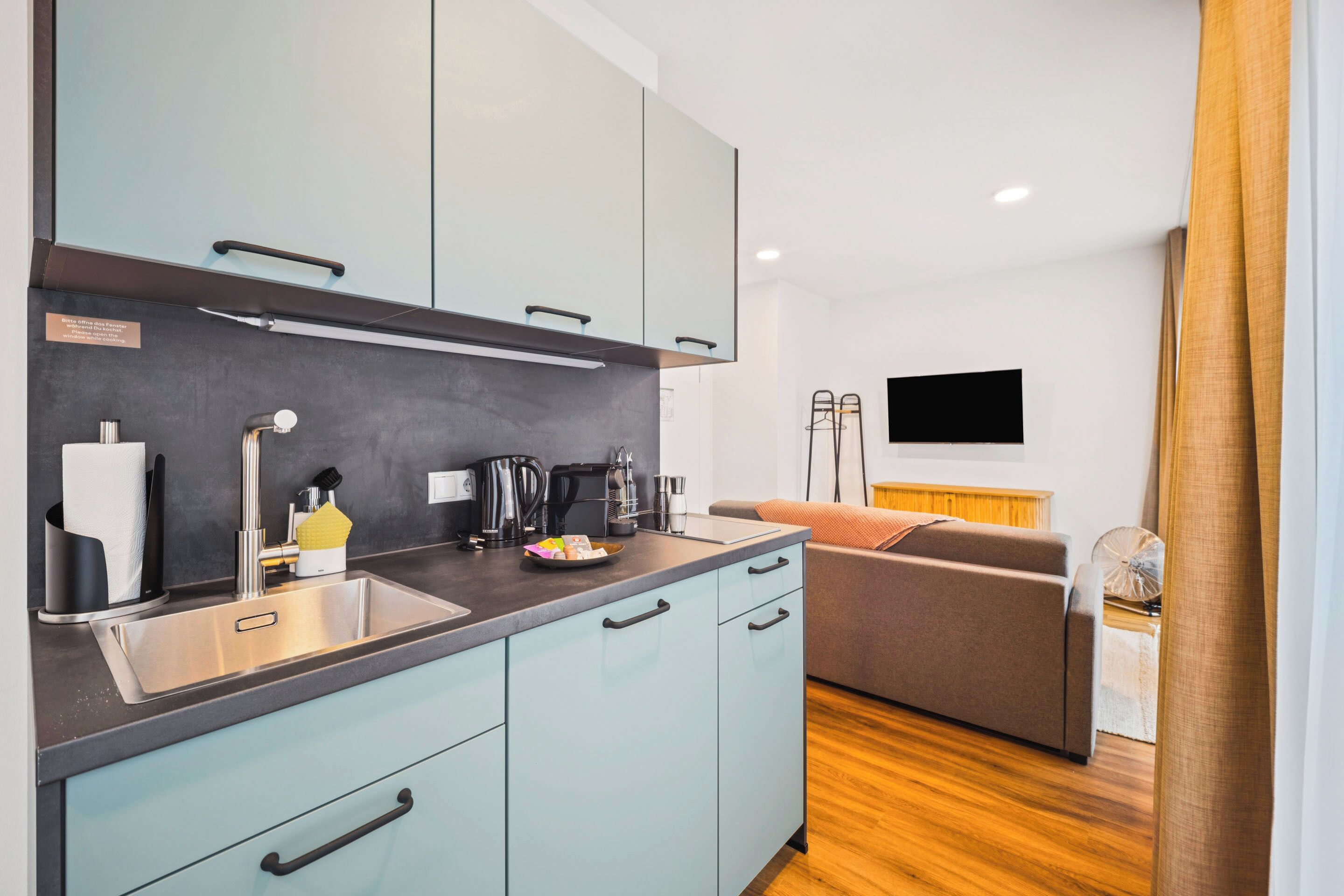 Una cocina moderna con armarios azul claro, un lavamanos de acero inoxidable y una cafetera. Hay un sofá y un televisor de pantalla plana en la sala de estar.