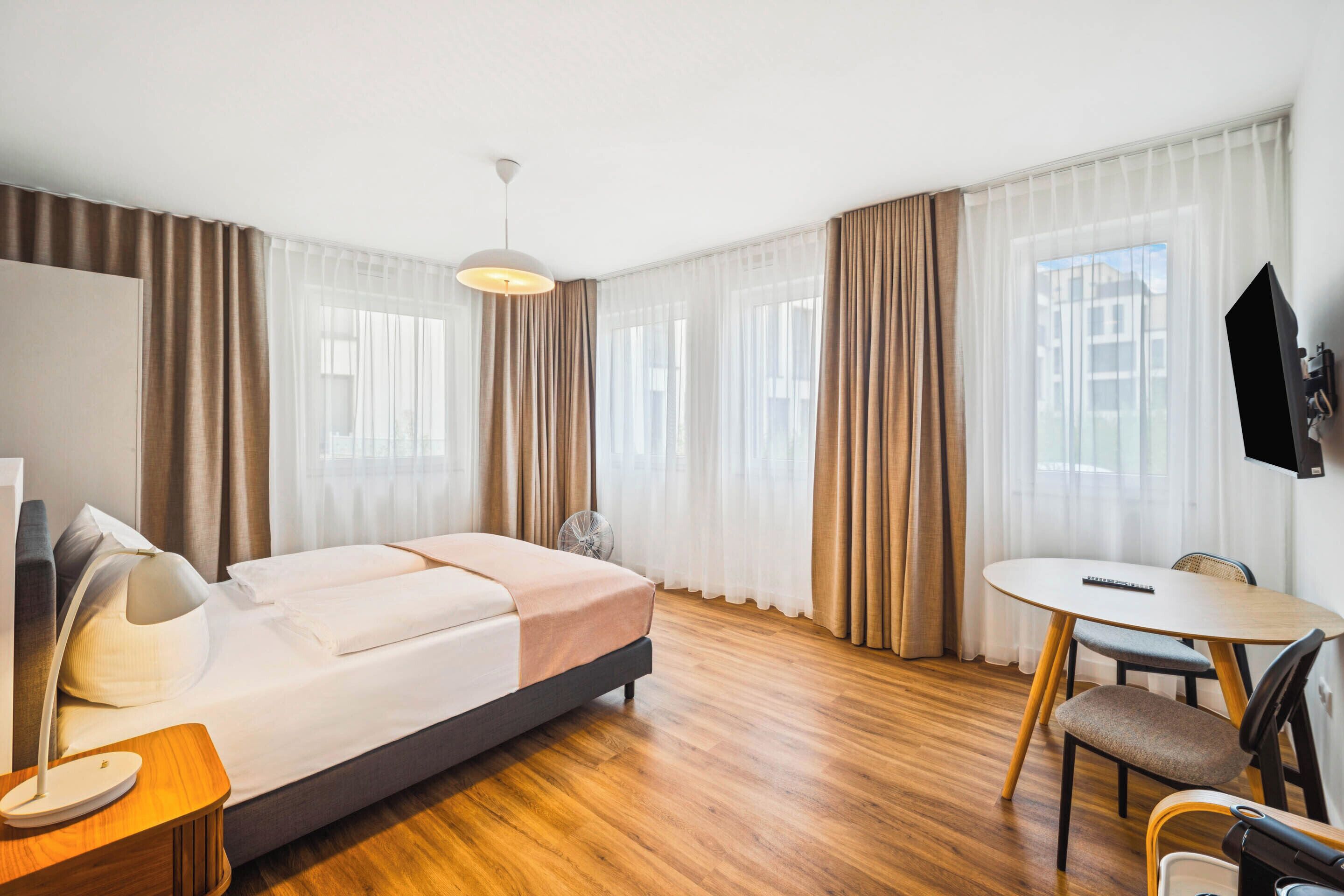 A modern hotel room with a bed, a desk, a chair, a fan, and a TV.