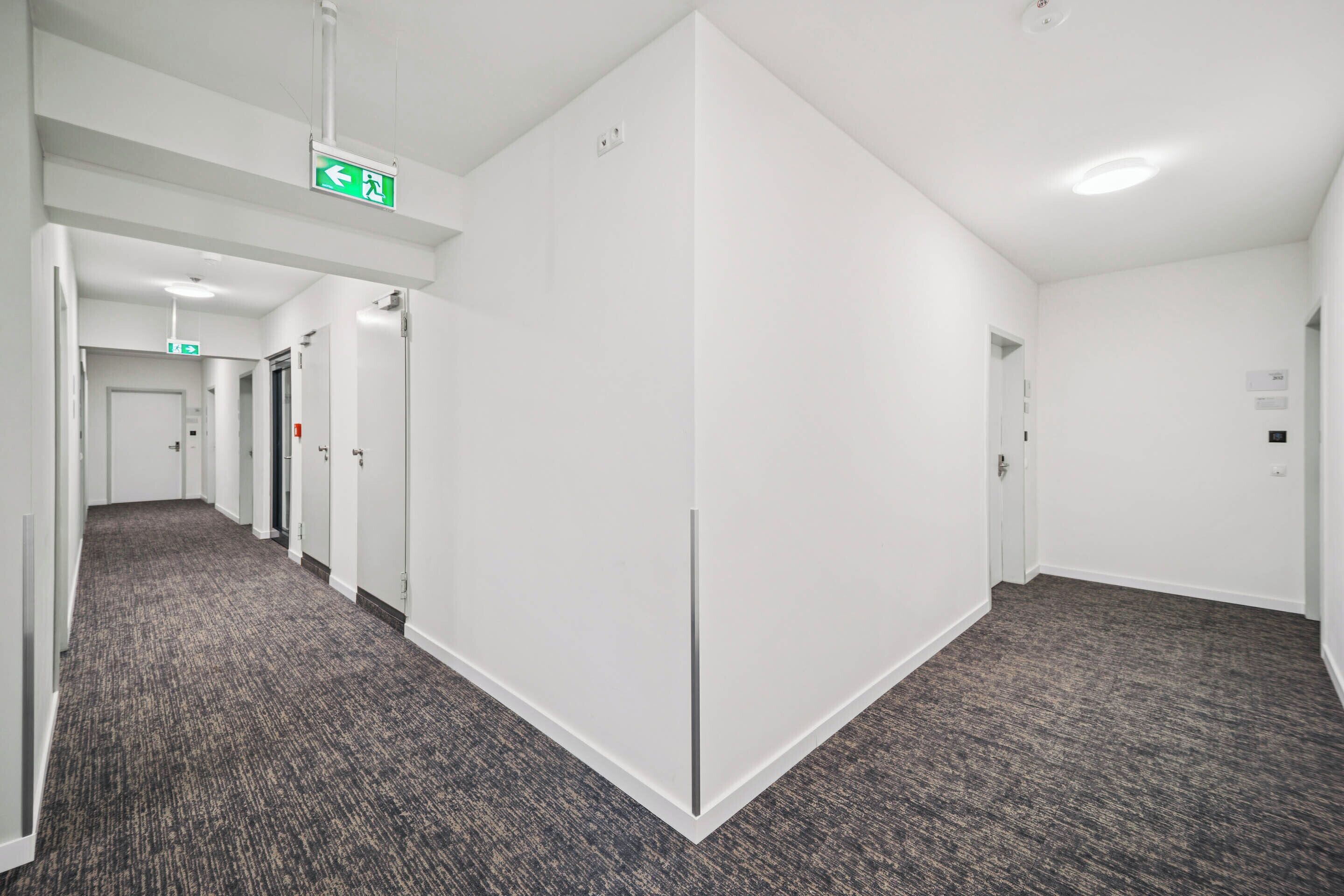 Un couloir d’hôtel aux murs blancs, au sol recouvert de moquette et dont les portes donnent accès aux chambres.