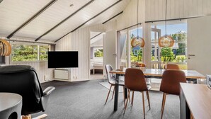 Une chambre moderne dotée d’une télévision à écran plat, d’une table à manger avec des chaises et de grandes fenêtres offrant une vue sur la verdure environnante.