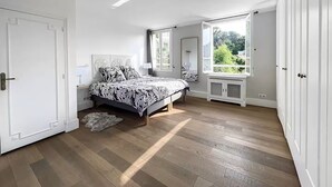 Une chambre à coucher comprenant un lit, une table de chevet, une commode et une grande fenêtre donnant sur un paysage verdoyant.