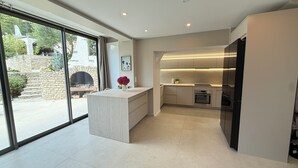 Une cuisine moderne dotée d’un îlot central, d’appareils électroménagers intégrés et offrant une vue sur une cheminée extérieure en pierre.