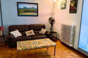 A living room with a brown leather sofa, patterned cushions, a quilted coffee table, a standing fan, a radiator, and a framed landscape painting on the wall. - Cozy apartment in historic center (Manziana)