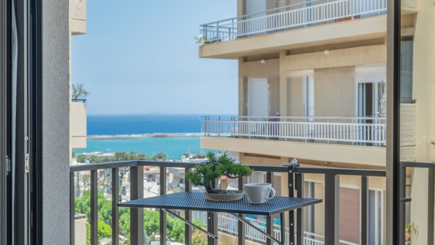 Un balcone con un tavolino, una pianta in vaso e una tazza. Il balcone si affaccia su un edificio con diversi piani e vista sul mare.