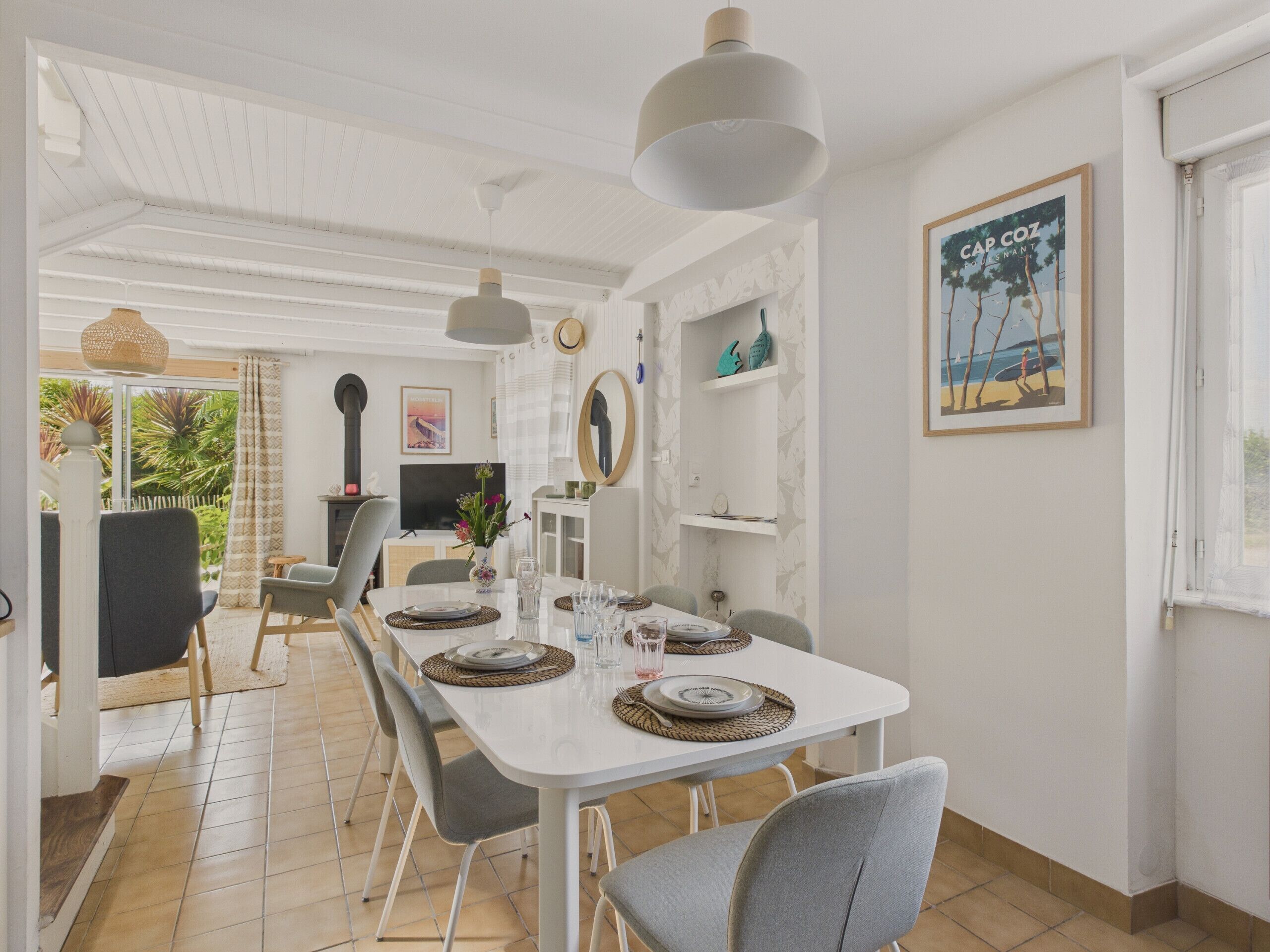A dining area with a white table set for four, surrounded by grey chairs. There is a large window on the right, a framed picture on the wall, and a ceiling fan above.