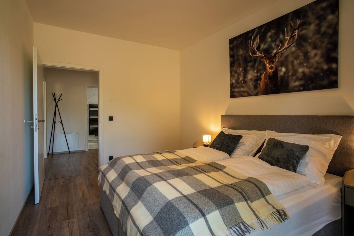 A bedroom with a large bed, a headboard, a nightstand, a lamp, and a painting of a deer on the wall.