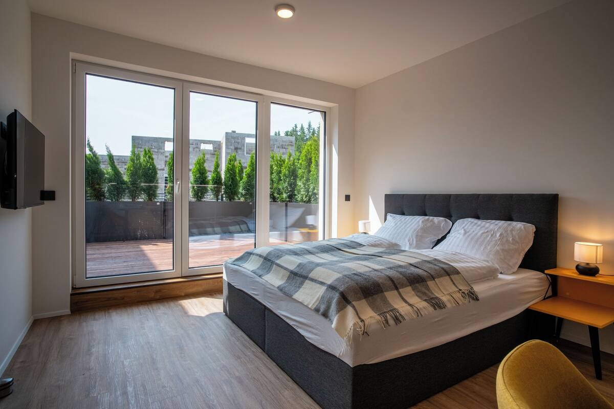 A modern bedroom with a large bed, a bedside table with a lamp, and a view through a sliding glass door to an outdoor balcony.