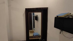 Een kamer met een deur, een spiegel, een klein krukje en een zwarte kast met een blauw voorwerp erop.