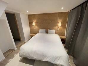 Une chambre avec une tête de lit en bois, deux lampes de chevet, un grand lit avec une literie blanche et un rideau foncé sur le côté droit.
