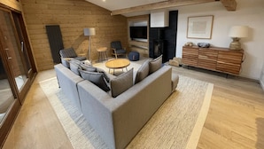 A modern loft-style room with a high ceiling, wooden beams, a skylight, a grey sofa, a small round table, a TV, and a wooden cabinet.