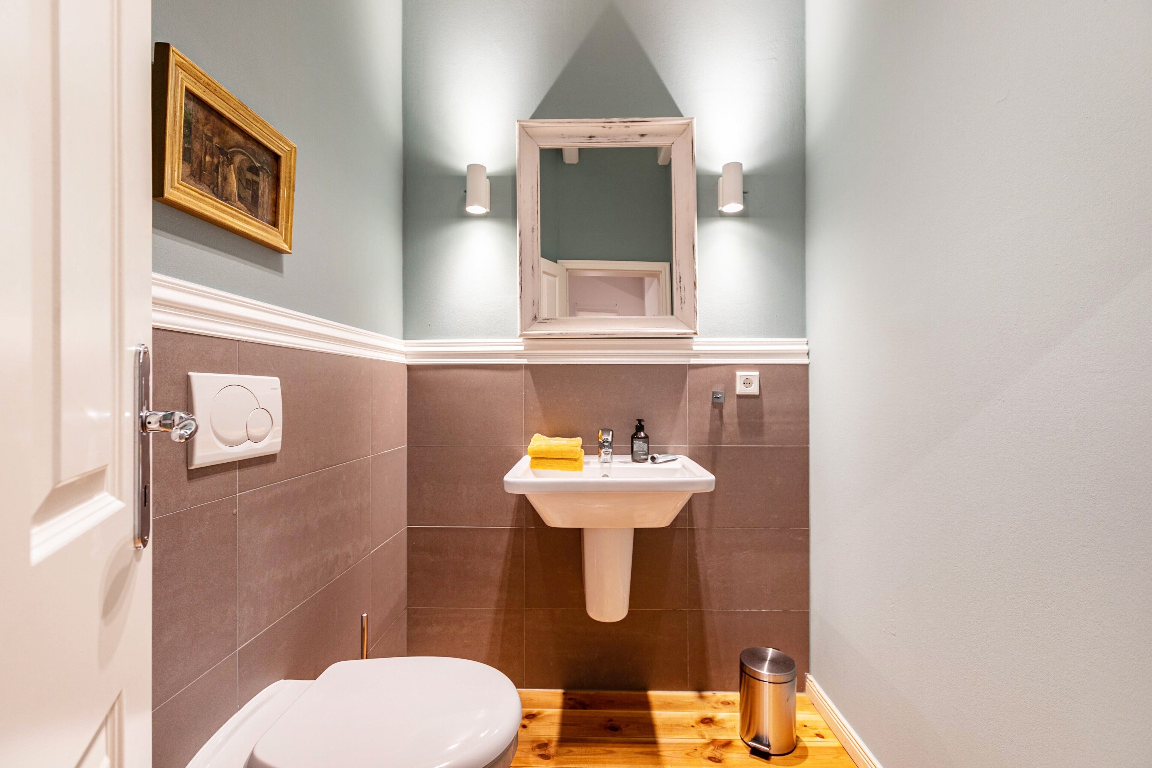 Een badkamer met een wit toilet, een wastafel met een gele handdoek, een lijstspiegel en een afbeelding aan de muur.