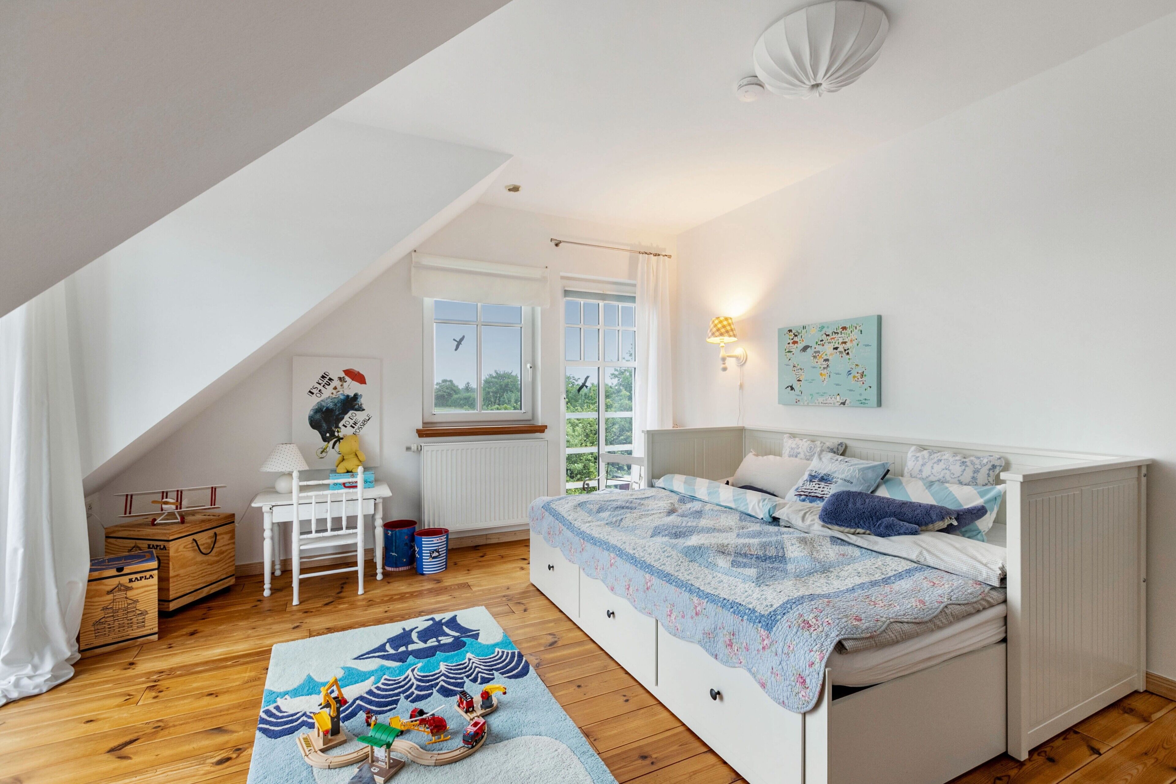 Une chambre avec un plafond en pente, un lit recouvert d’une courtepointe, un bureau avec une lampe et une fenêtre avec des rideaux.