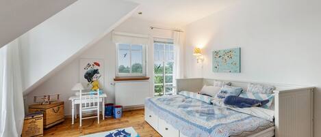 Une chambre avec un plafond en pente, un lit recouvert d’une courtepointe, un bureau avec une lampe et une fenêtre avec des rideaux.