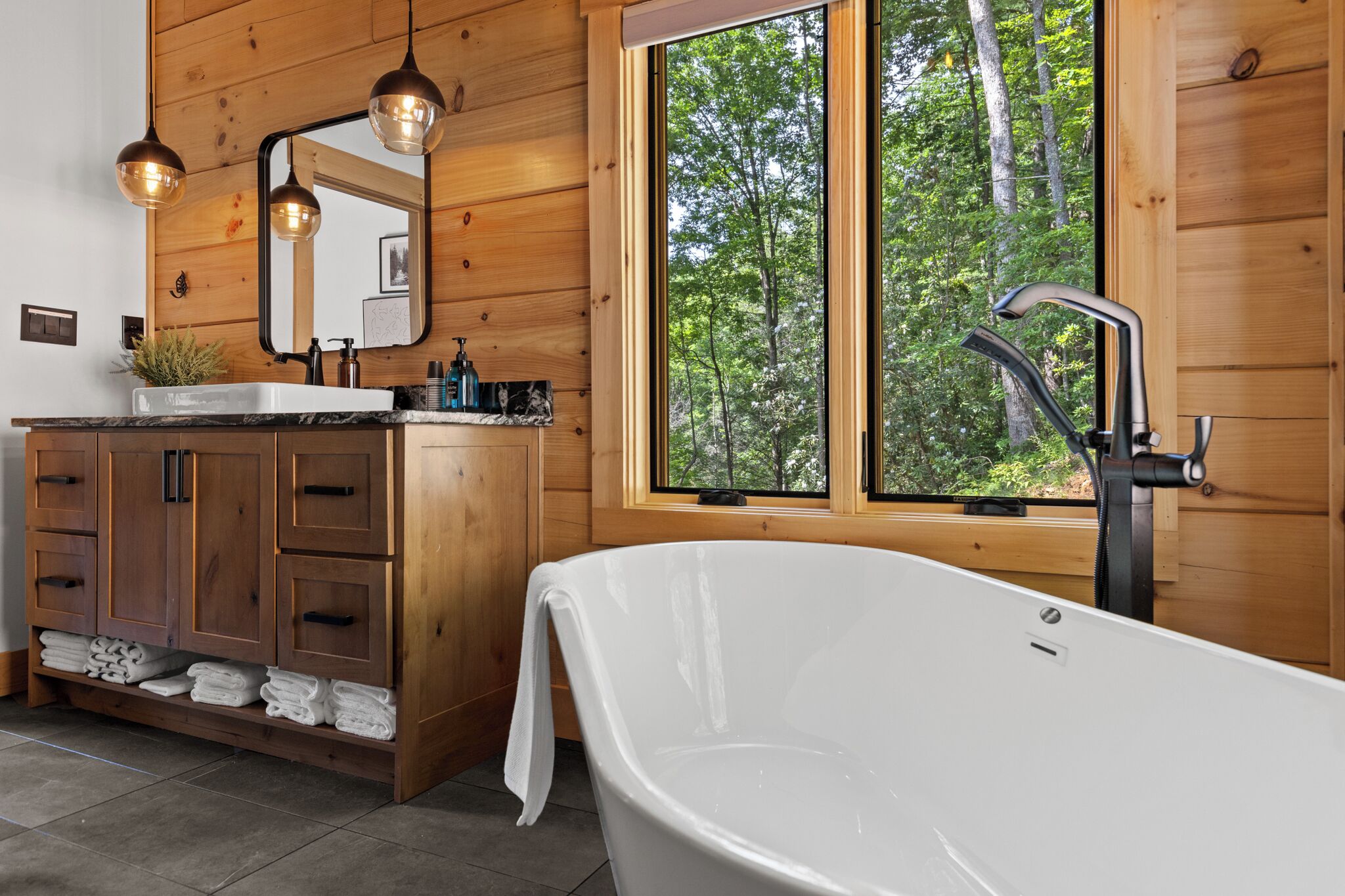 Une salle de bain rustique, avec une cabine en bois, une grande baignoire, des meubles en bois, un lavabo avec un miroir et une fenêtre donnant sur la forêt.