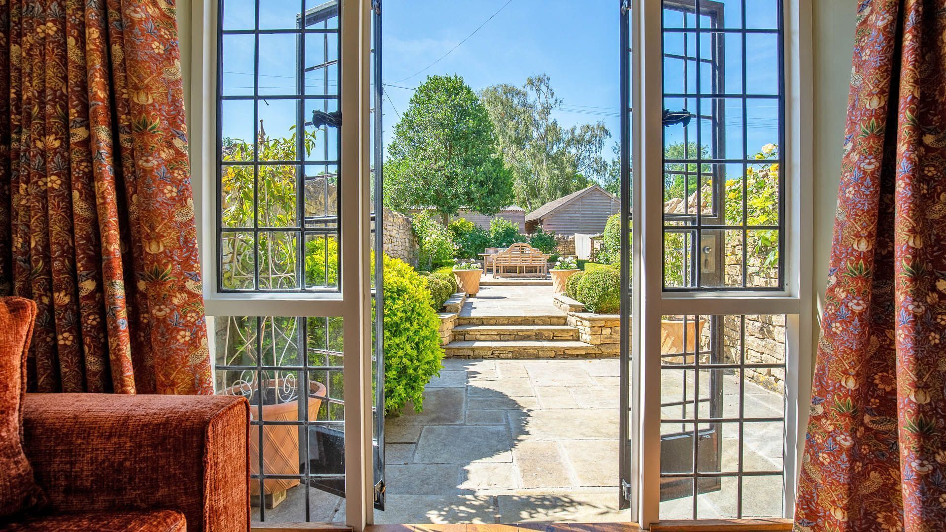 A view through a glass door leading to a stone pathway, a wooden bench, and a garden with various plants.