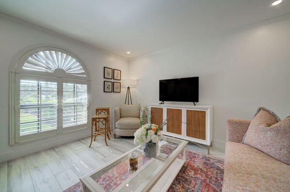 A living room with a large arched window, a sofa, a television, a cabinet, and a coffee table with a vase of flowers. - LeMalo Vacation Rental: Stylish 1-BR Condo Near 5th Ave S & Gulf Beaches Coastal Vibes, King Bed, and Private Outdoor Space. Perfect Location for Dining, Shopping and Exploring Naples! (Naples)