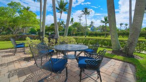 A patio with a round metal table and chairs, surrounded by palm trees and a well-maintained garden. - LeMalo Vacation Rental: Stylish 1-BR Condo Near 5th Ave S & Gulf Beaches Coastal Vibes, King Bed, and Private Outdoor Space. Perfect Location for Dining, Shopping and Exploring Naples! (Naples)