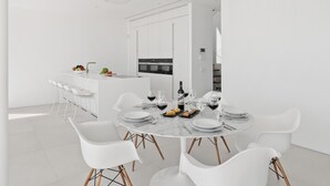 Uma sala de jantar moderna e minimalista com uma mesa redonda de mármore, cadeiras brancas e cozinha ao fundo.