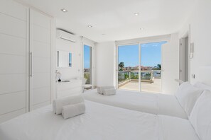 A modern bedroom with a large bed, a built-in wardrobe, a bathroom with a sink and mirror, and a balcony with a view of buildings and palm trees. - Villa El Duque Privé (Adeje)