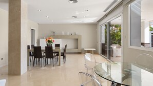 Et moderne spiseområde med et glasbord og stole, et hvidt spisebord med stole, samt et køkken med skabe og bordplade.