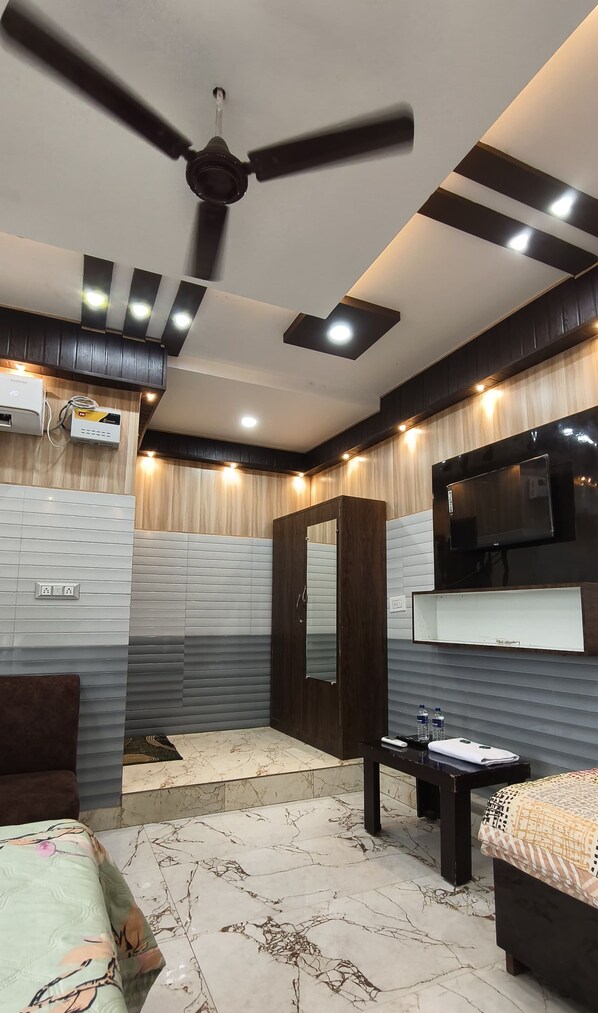 A modern bedroom with a ceiling fan, a TV mounted on the wall, a bed with a patterned bedspread, and a bedside table with a remote control and water bottles. - Hotel Samiksha Inn (Ayodhya)