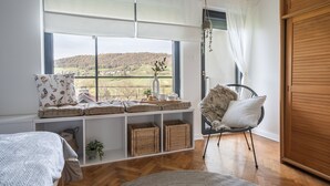 Un dormitorio con un ventanal grande, piso de madera, cama, una silla, repisas con cestas y una planta colgante.