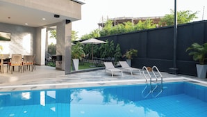 Une piscine entourée d’un mur sombre et de verdure, avec des chaises longues et une table à proximité.